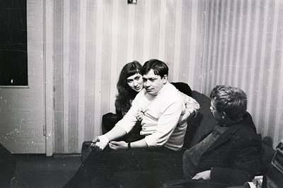 A candid black and white image depicts three young adults seated in armchairs. A man with a sweater and short hair is embrace...