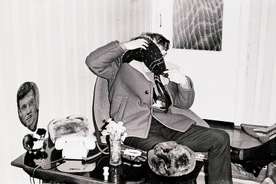 A man in a dark overcoat and scarf sits slumped on a chair, rubbing his forehead. The room features a patterned wallpaper and...