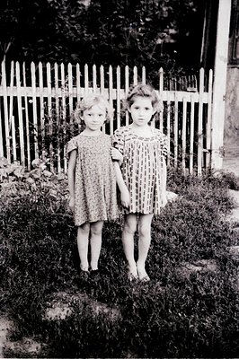 Two young girls, barefoot and wearing vintage floral print dresses, stand side-by-side in a garden. A white picket fence and ...