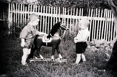 Two young boys stand near a wooden rocking horse in a grassy yard, captured in a vintage black and white photograph. The boys...