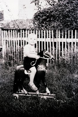 A young child sits on a dark, vintage rocking horse in a grassy backyard. The child wears a short-sleeved shirt and sandals. ...