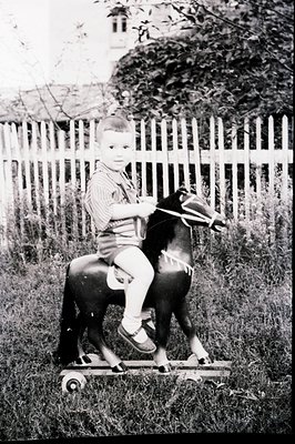 A young child rides a rocking horse outdoors, seated on a wooden platform with wheels. The scene features a white picket fenc...