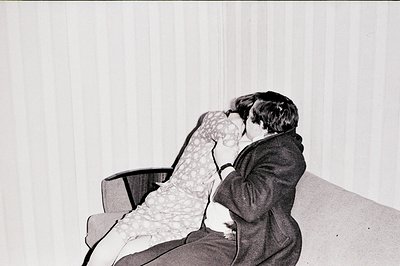 A couple embraces in a domestic interior, the man in a dark suit, woman in a patterned dress seated on an armchair. Stark, te...
