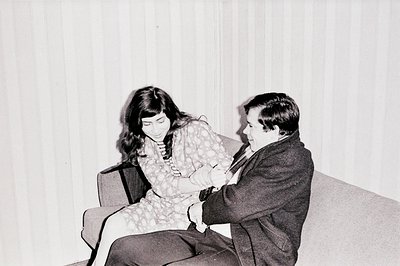 A young couple seated on a textured, patterned sofa, engaged in conversation. The woman wears a floral print dress, while the...