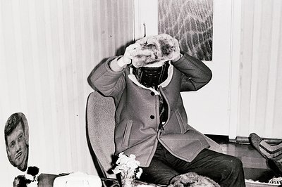 Man sits in a cushioned chair, partially obscured by a large, furry hat. Visible details include a textured wool coat and pat...