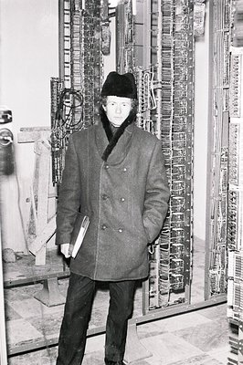 Man in a double-breasted overcoat and fur hat stands before a complex electrical panel. Likely a mid-20th century workplace, ...
