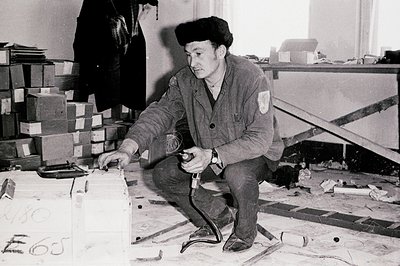 A man in a dark beret and workwear kneels, operating a rivet gun near stacked boxes. The interior space appears industrial or...