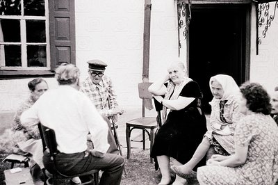 Group portrait captures a moment of rest or conversation outdoors. Five individuals are seated on mismatched chairs in what a...