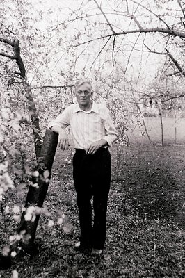 Portrait of a man leaning casually against a blossoming tree. He wears a collared shirt, dark trousers, and appears to be out...