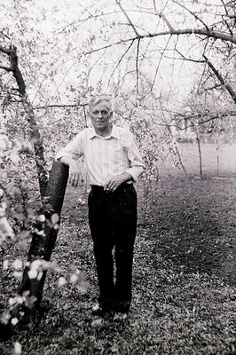 A gentleman in a collared shirt and trousers poses leaning against a tree with blossoms. Appears to be an outdoor portrait, l...