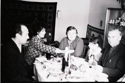 A family gathers around a laden table covered with bowls of food. Black and white photography, likely 1960s or 70s. Interior ...