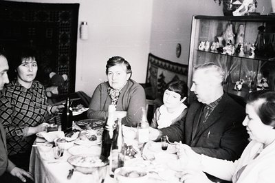 A family gathers around a table laden with food, likely a holiday meal. Interior shows a heavily-decorated display case with ...