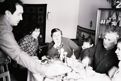 A family gathers around a laden table, likely celebrating a special occasion. Interior scene shows a traditional decorative s...