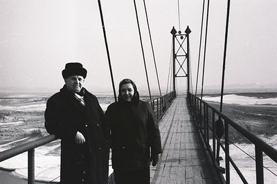 A couple stands proudly on a weathered suspension bridge, overlooking a windswept coastal landscape. The man wears a hat & lo...