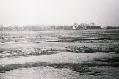 A wide, frozen landscape under a heavy, overcast sky. Textured, uneven ice covers the foreground, transitioning to a distant ...