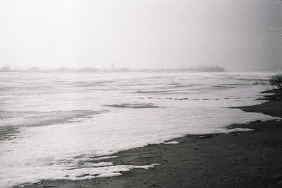 A wide, grayscale view of a frozen shoreline with low-lying mist obscuring distant land. Dark sand contrasts with the icy, te...