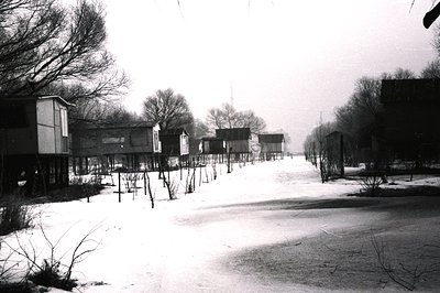 A stark, monochrome view of a village or settlement in winter. Simple, elevated wooden structures line a snow-covered road. P...