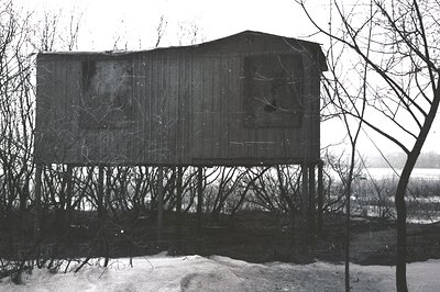 Weathered, elevated structure with a peaked roof sits above a snowy landscape. The building’s wood siding appears aged and sh...