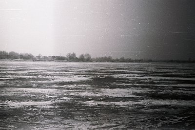 Frozen expanse of water, likely a lake or reservoir, stretches toward a distant treeline and village. Heavily textured surfac...