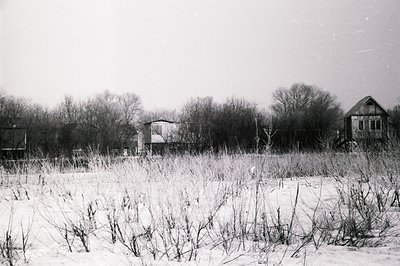 Bleak winter scene: snow-covered meadow transitions to a line of weathered, wooden buildings and bare trees. Likely rural set...