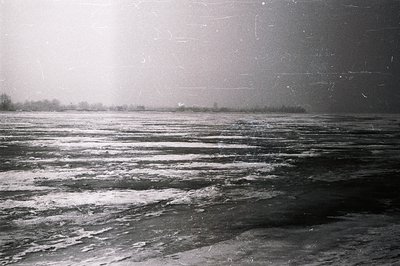 A monochromatic landscape depicts a frozen body of water, likely a lake or river, extending to a distant, hazy shoreline. The...