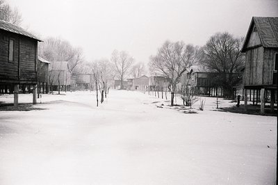 A snow-covered village, likely a rural or logging settlement, with wooden buildings on stilts. Bare trees line the unpaved ro...