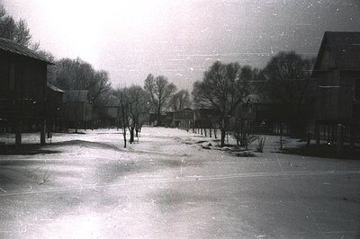 A monochrome image captures a snow-covered village scene. Log cabins with dark roofs line a central, snow-packed path. Sparse...