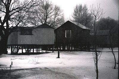 Rustic, raised cabins sit on wooden stilts amidst a snowy landscape. A solitary wooden post stands prominently in the foregro...