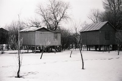 Two elevated, vernacular structures stand in a snow-covered field. The buildings feature simple gable roofs and appear to be ...