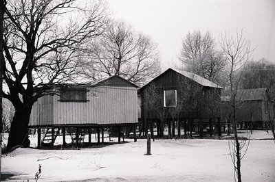 Elevated, vernacular structures on piers define this stark winter scene. Likely rural dwellings, the simple wood architecture...