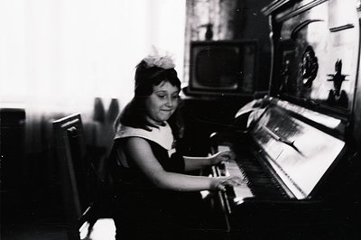 A young girl seated at an upright piano, appears to be playing. Visible in the background is a vintage television set and fra...
