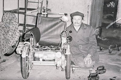 A seated man in workwear poses alongside a vintage three-wheeled vehicle, likely a prototype or restoration project. The sett...
