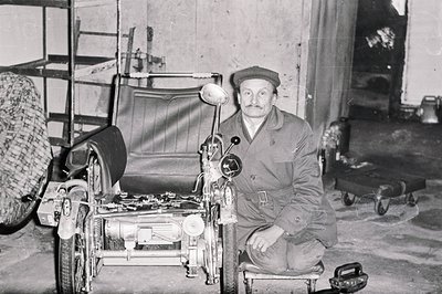A man in work overalls and cap poses proudly beside a custom-built, three-wheeled vehicle resembling a miniature car. The int...