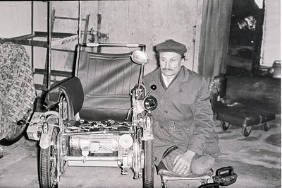 A man in a cap and work attire sits beside a unique, possibly custom-built, three-wheeled vehicle. The image shows a workshop...