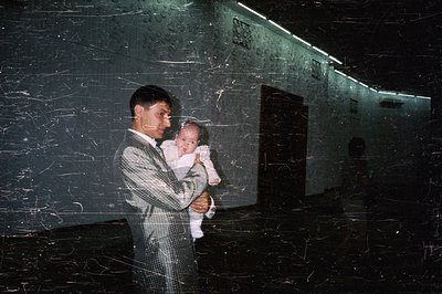 Man in a patterned suit holds a baby, likely indoors, within a stark, modernist architectural space. Visible surface texture ...