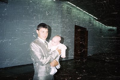 A man in a gray checkered suit holds a baby wrapped in white, standing in a stark, modernist interior. The space features dar...