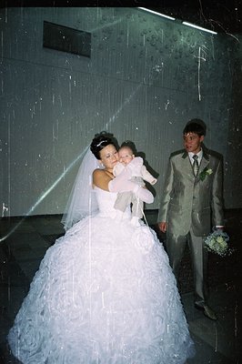 A bride in a voluminous, heavily-layered, ruffled wedding gown cradles an infant; a groom in a patterned suit stands beside t...