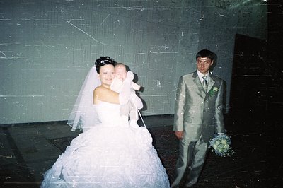 A bride in a voluminous, strapless gown holds a baby bundled in white, while a groom in a textured suit stands beside them. L...