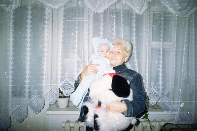A woman with styled hair holds a baby wearing a blue bonnet indoors, near a window draped with lace curtains. She wears a dar...