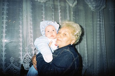 A woman with short, blonde hair cradles an infant in a blue onesie. The photo, likely taken indoors, features patterned, shee...