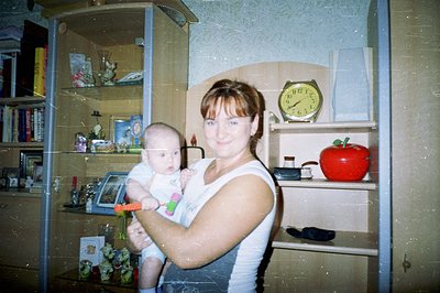 A woman cradles a baby while standing in a room with decorative shelves. The shelves hold a clock, a red apple, picture frame...