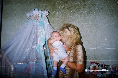 A blonde woman holds an infant in patterned clothing and blue socks. A lace-trimmed canopy hangs behind them. Likely a family...