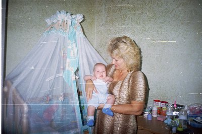 Woman in shimmering gown cradling a baby, posed in front of a vintage baby crib draped with lace netting. Wallpaper features ...