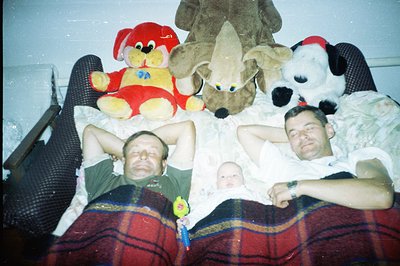 A humorous snapshot, likely family, featuring two men playfully reclining behind a sleeping infant bundled in white. Large pl...