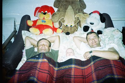 Three men relax on a large bed covered in a plaid blanket, flanked by oversized plush animal toys. A baby rests between two a...