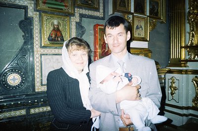 A couple holds a baby, likely during a christening ceremony within an Eastern Orthodox church. The background features numero...