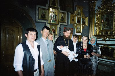 Group portrait within an ornate Orthodox church; a priest holds an infant swaddled in white. Six individuals, including the p...
