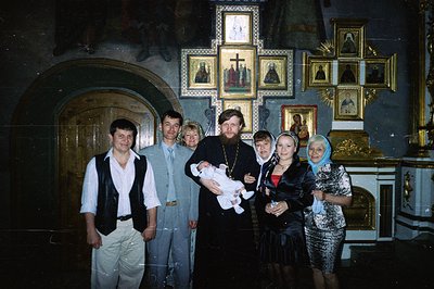 A formal portrait within a church, likely depicting a christening ceremony. Six individuals are grouped around a bearded Orth...
