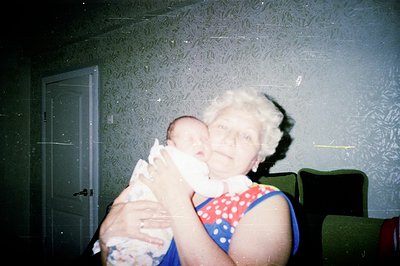 An elderly woman with styled white hair holds a swaddled newborn infant. Interior scene features patterned wallpaper and a da...
