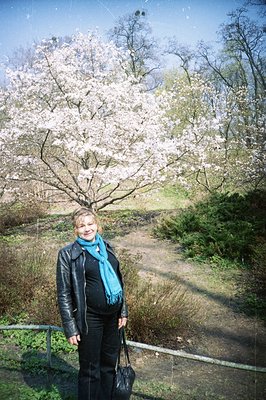 A woman in a black leather jacket and scarf poses beside a flowering tree in full bloom. Likely mid-1990s color photography, ...